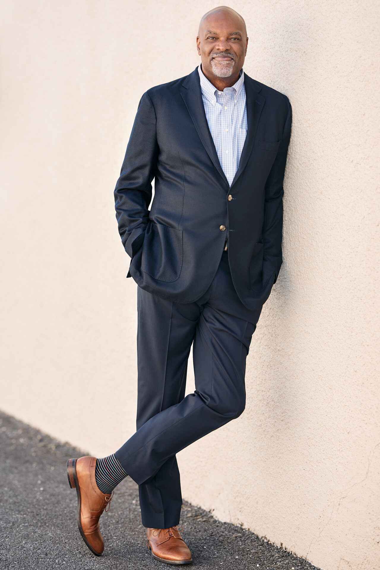 A man in suit leaning against wall with hands in pockets.